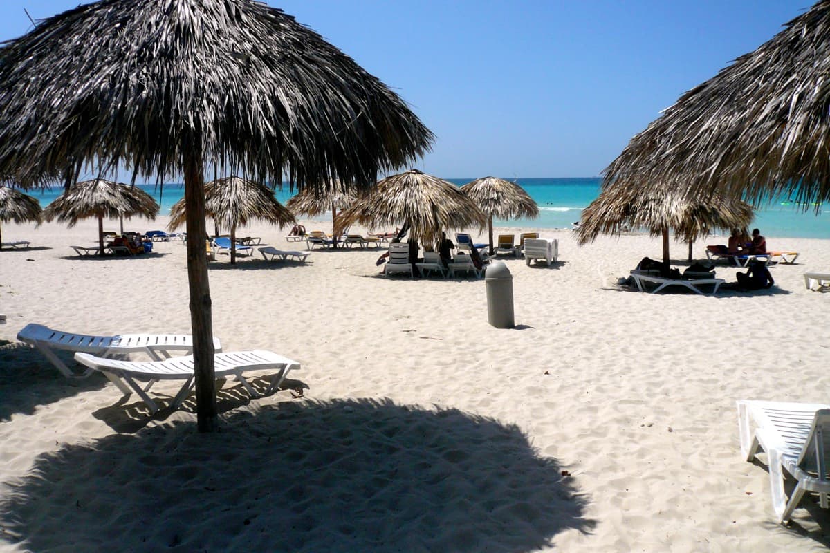 A long empty white-sand beach with turquoise sea and green palm trees in Cuba
